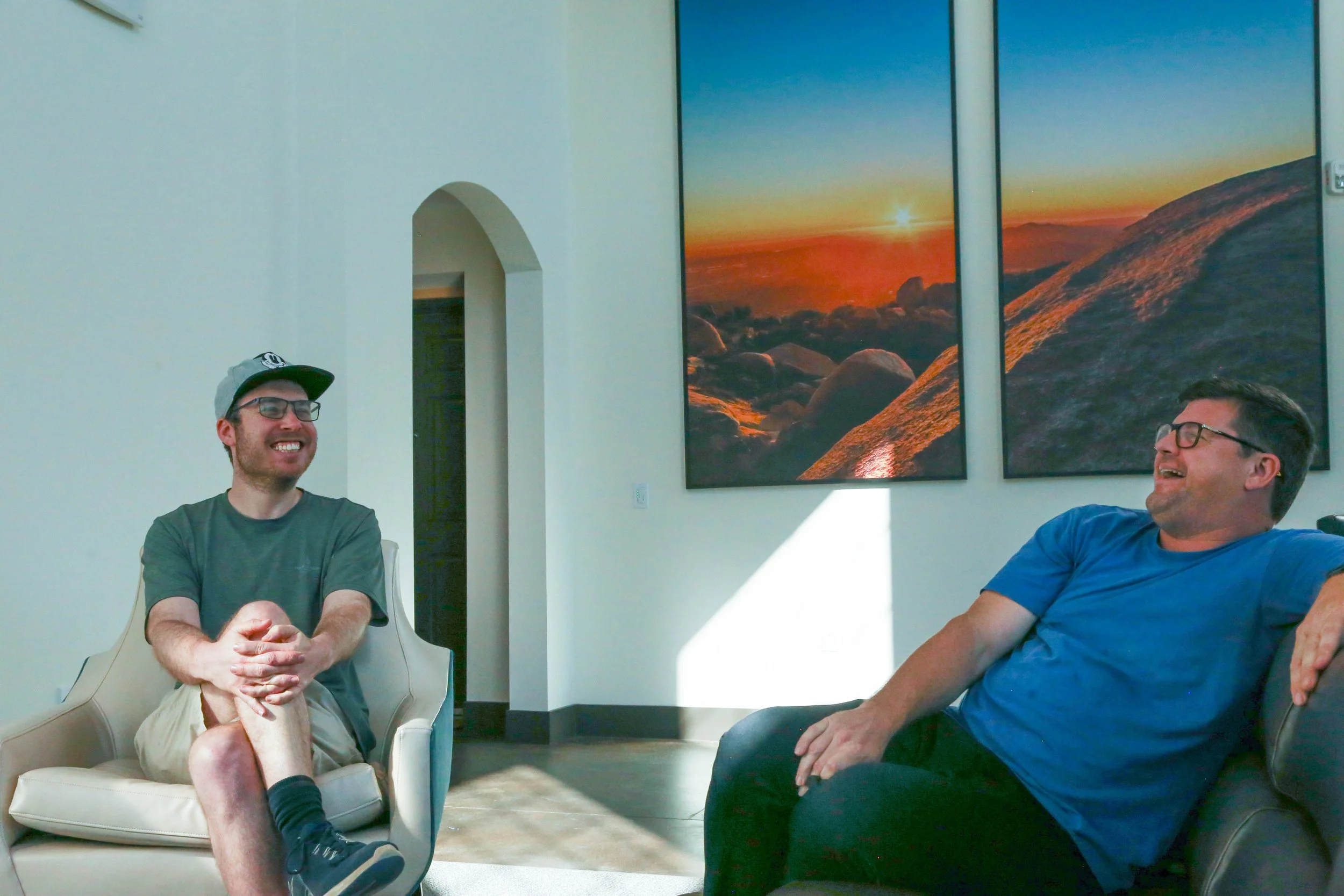 Two men are sitting and laughing in a living room with white walls and large landscape photographs of a sunset over rocks on the wall behind them.