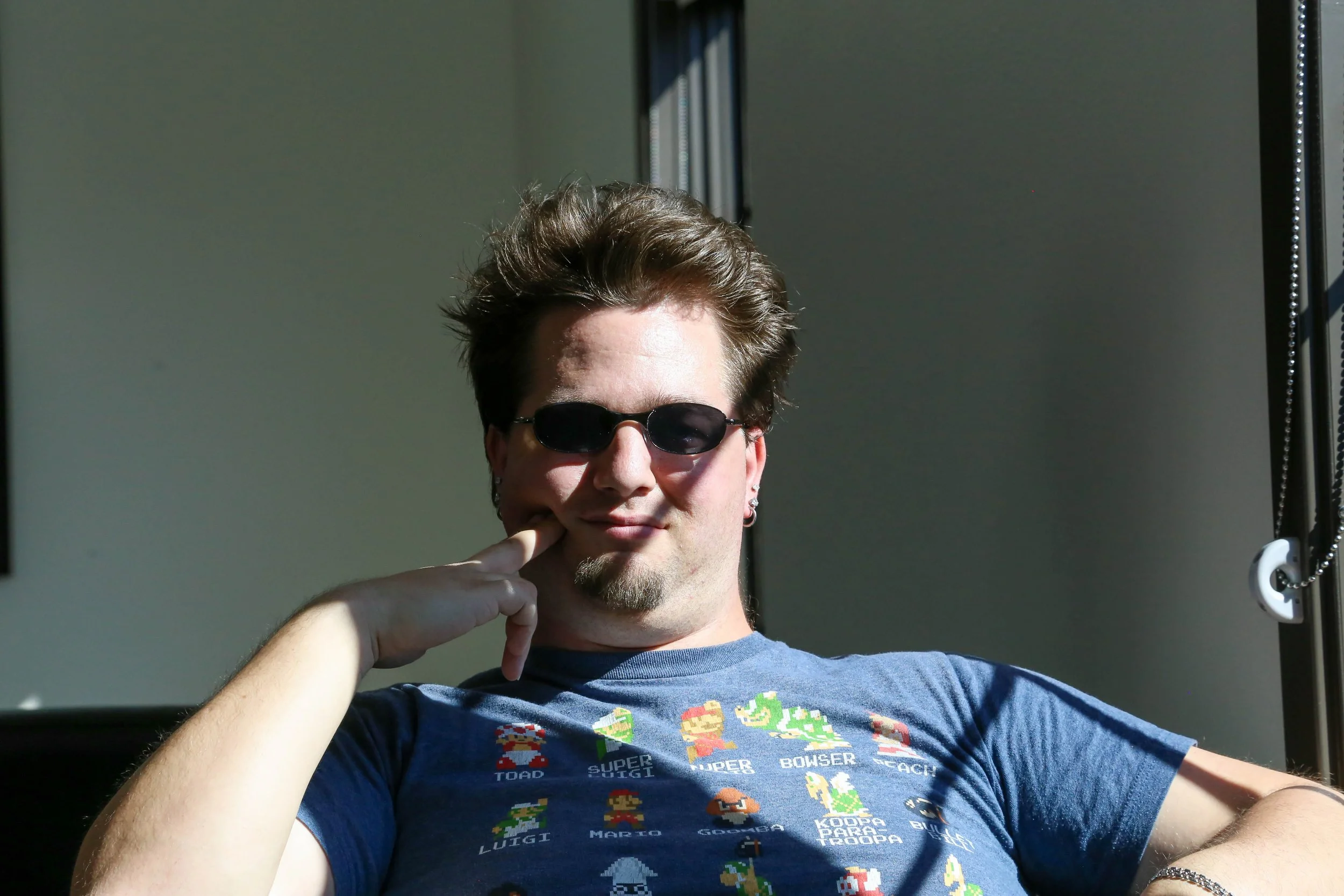 A young man with brown hair, wearing sunglasses, a blue Super Mario T-shirt, and earrings, sitting near a window with sunlight on his face and a neutral expression.