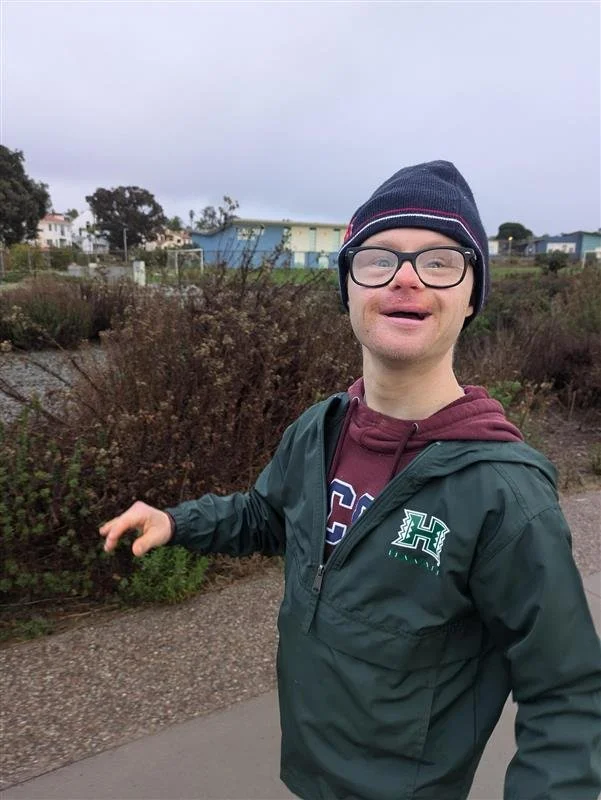 Person with Down syndrome wearing glasses, a navy beanie, a green jacket, and a maroon hoodie walking outdoors on a cloudy day.