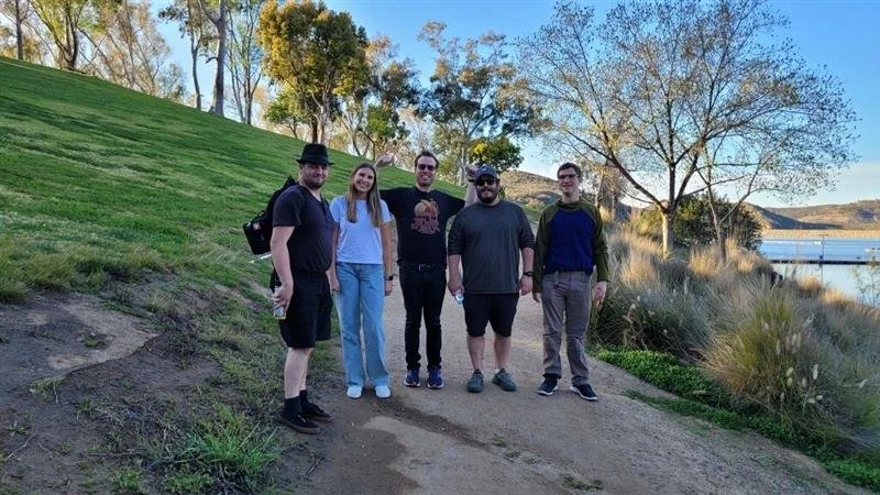 Five people walking outdoors on a dirt path next to a grassy slope and a body of water, with trees and hills in the background during daytime.