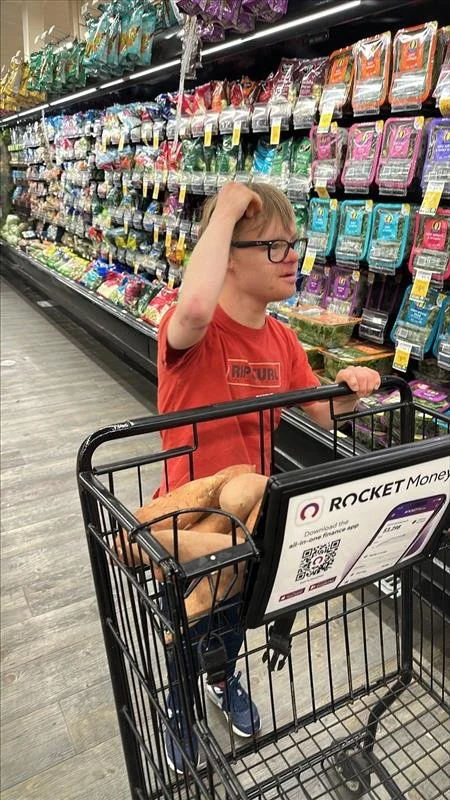 A young boy with glasses and a red Rip Curl T-shirt standing in a grocery store aisle with a shopping cart filled with potatoes and bread, holding the cart handle while scratching his head, surrounded by shelves of snack bags.
