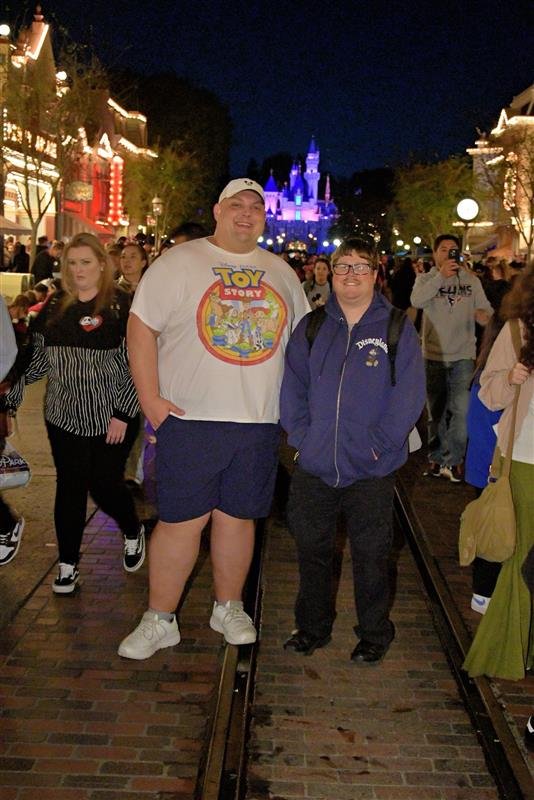 Two people stand on a brick street at a theme park at night, with a lit castle in the background. One is wearing a Toy Story shirt and shorts, the other is wearing a blue jacket and glasses.