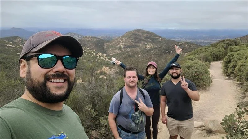 Four friends hiking on a trail in a mountainous area with green bushes and distant peaks, taking a group selfie.