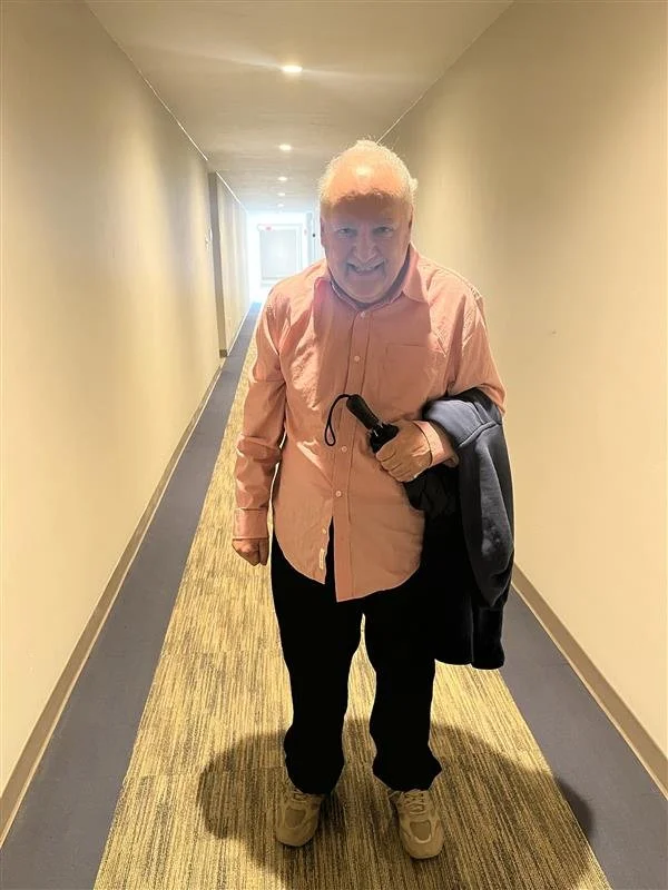 An elderly man walking in a hallway, smiling, wearing a pink button-up shirt, holding a black umbrella and a dark jacket, with beige shoes, and a gray sweater draped over his arm.