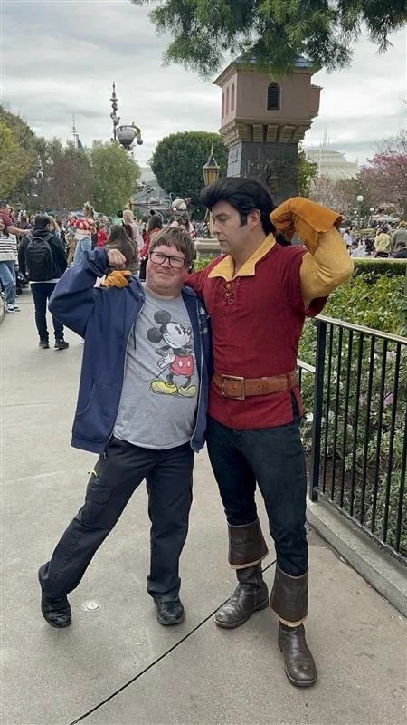 A young man posing with a costumed character dressed as Gaston from Disney's Beauty and the Beast, both making flexing arm poses in an amusement park with other visitors in the background.