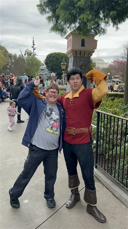 A man in casual clothes with a Mickey Mouse shirt standing next to a person dressed as Gaston from Disney's Beauty and the Beast at a theme park, with other visitors and rides visible in the background.