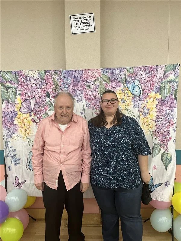 An elderly man and a young woman standing side by side in front of a spring-themed backdrop with butterflies and flowers, surrounded by colorful balloons.
