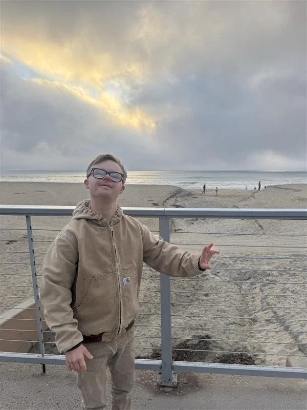 A young boy with glasses and blonde hair smiling while standing next to a metal railing at the beach during sunset, with the ocean in the background and a cloudy sky.