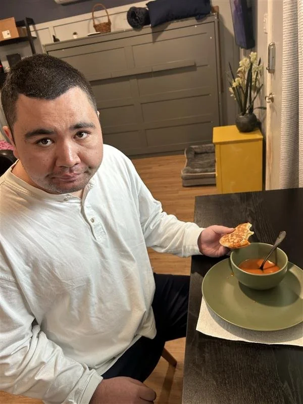 Man sitting at a dining table holding a piece of bread, with a bowl of soup and a spoon on a plate in front of him.