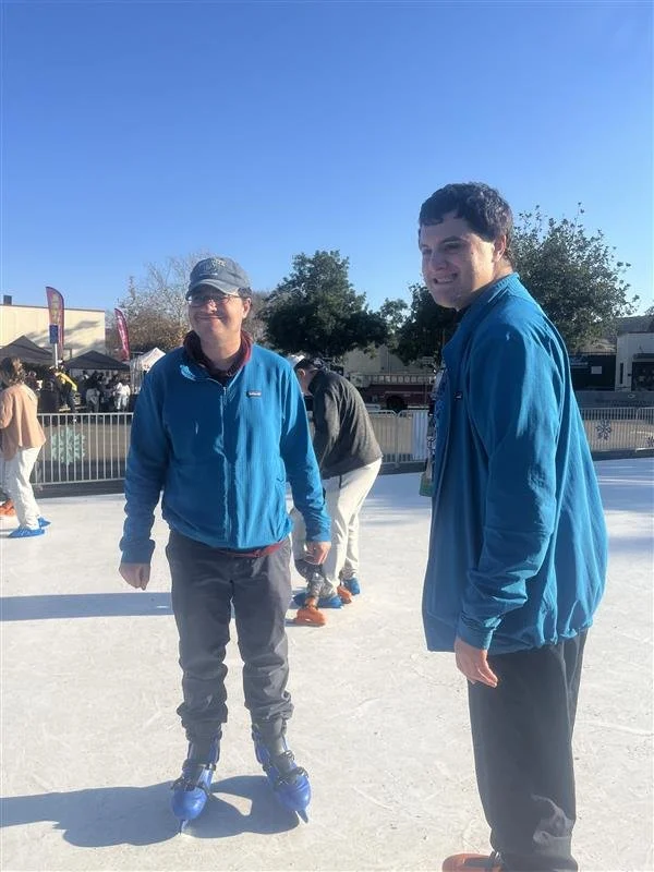Two people ice skating outdoors on a sunny day, wearing blue jackets and surrounded by other skaters and trees.