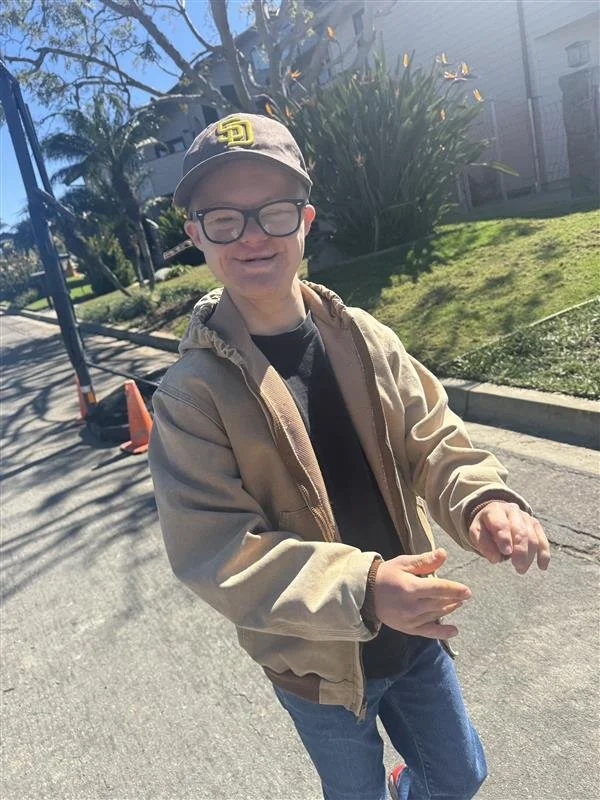 A young boy wearing glasses, a baseball cap, a beige jacket, and jeans standing on a sidewalk in a sunny neighborhood with trees and houses in the background.