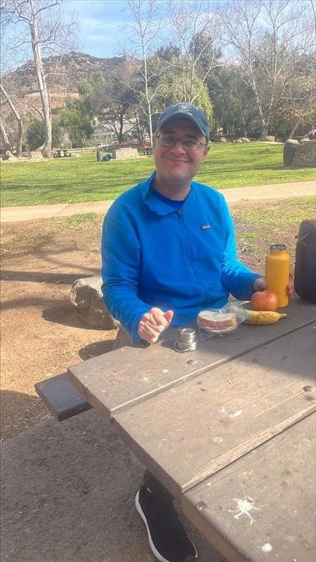 A man in a blue jacket and baseball cap sitting at a park picnic table with food and drinks, smiling at the camera.