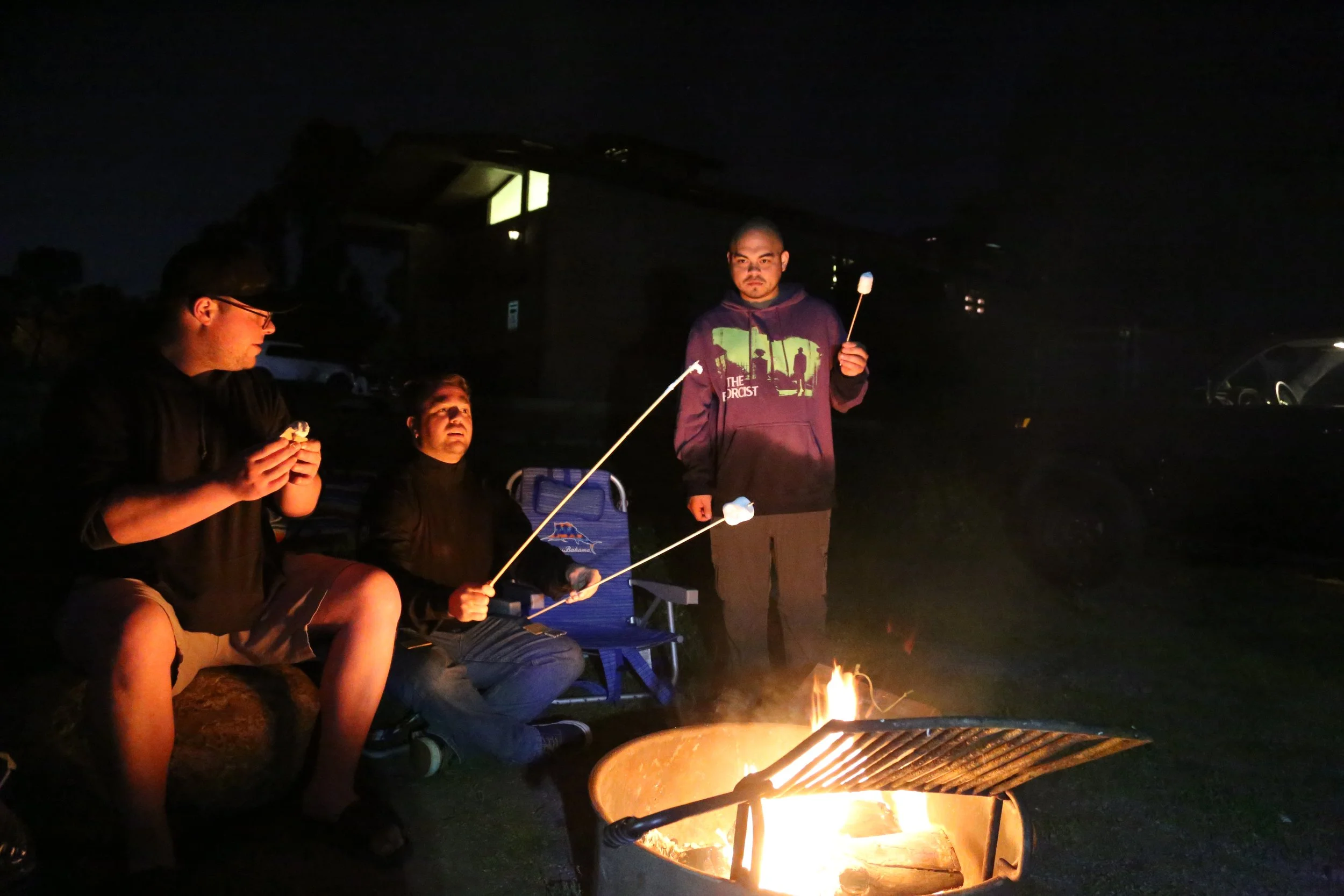 Three friends sitting and standing around a campfire at night, roasting marshmallows. One person is sitting on a rock, another on a camping chair, and the third is standing holding a marshmallow on a stick. The scene is outdoors with parked cars and 
