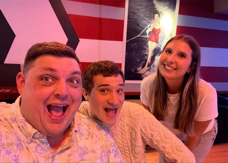 Three people, two men and one woman, smiling and posing for a selfie in a room with colorful walls and a painting of a woman water skiing in the background.