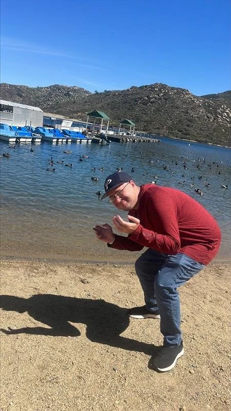 Man in red jacket and baseball cap crouching on sandy beach near lake, with ducks swimming in water and mountains in background.