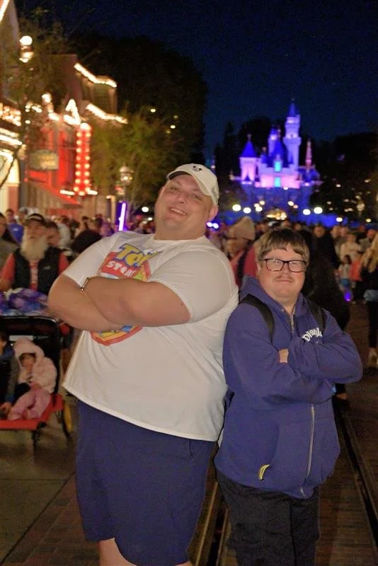 Two men standing together at night in front of a Disney castle illuminated with purple and blue lights. The man on the left is smiling with arms crossed, wearing a white cap and a T-shirt with a superhero logo. The man on the right has glasses, arms 