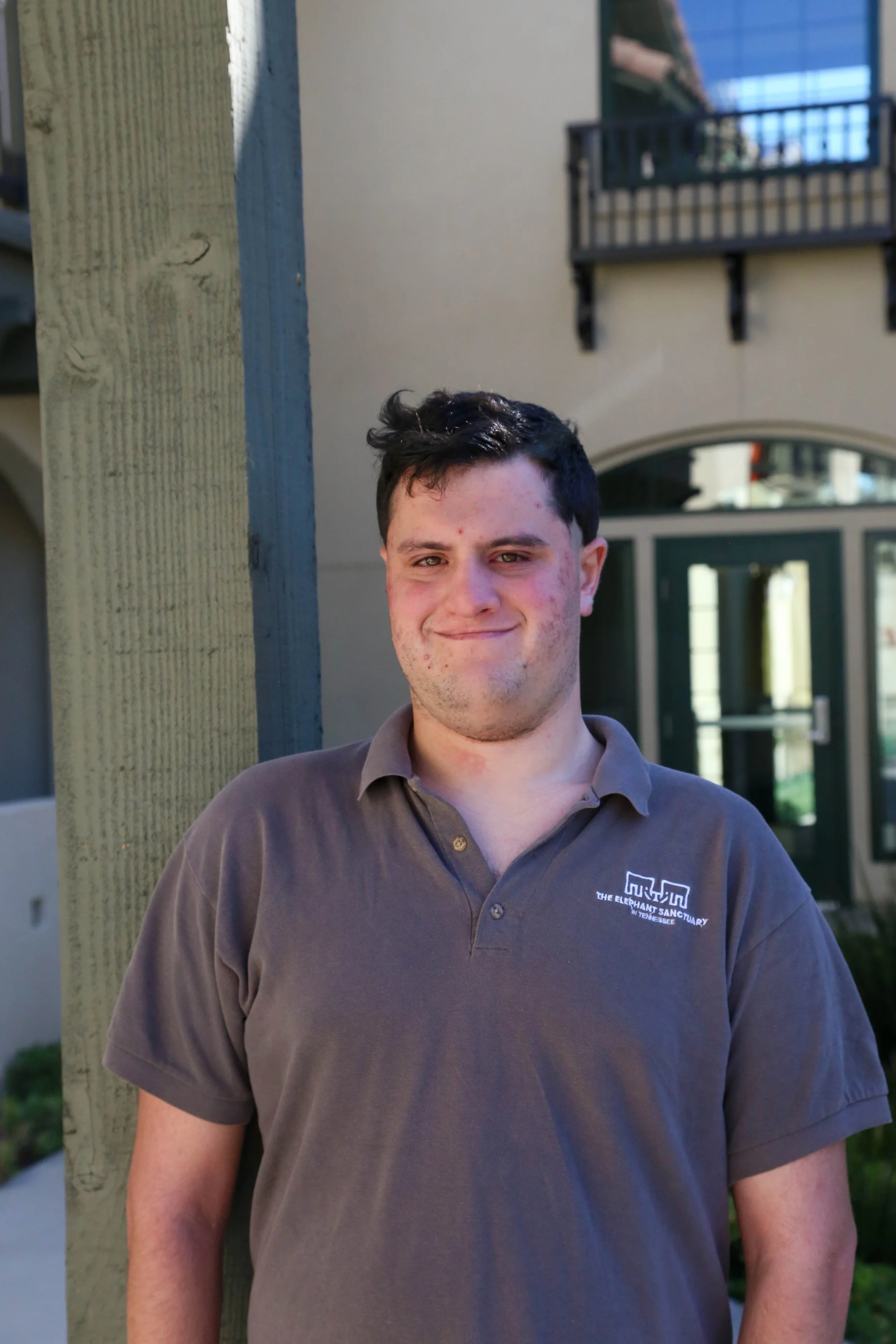 A young man with short dark hair and light skin, standing outdoors in front of a building with a balcony. He is wearing a gray polo shirt with a logo on the chest. The background includes a green door and window, and he is standing beside a wooden po