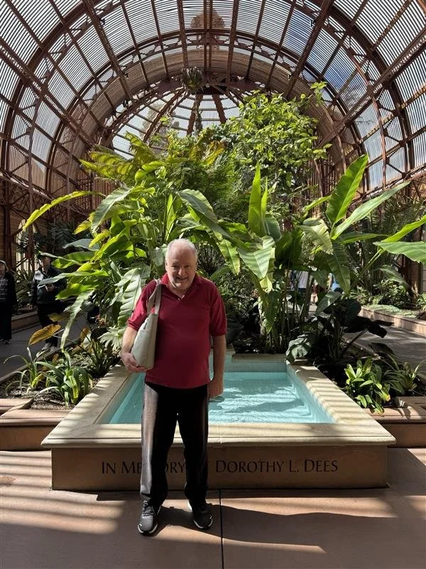 An elderly man standing in front of a small water feature inside a large glass greenhouse filled with lush green tropical plants.