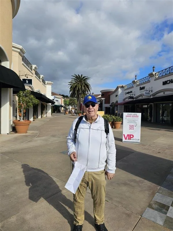 A man stands on a pedestrian shopping street, wearing a white jacket, khaki pants, sunglasses, a blue cap, and carrying a backpack and shopping bag. There are stores and a palm tree in the background under a partly cloudy sky.