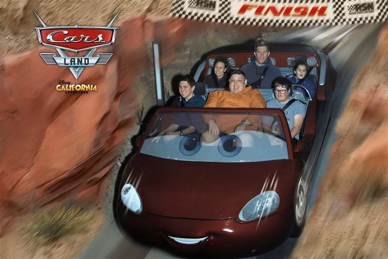 A group of six children and a teen riding on a 'Cars Land' roller coaster themed ride at California Adventure park. The coaster is designed to look like a shiny red race car with animated eyes and a smiling mouth, going through a desert canyon settin