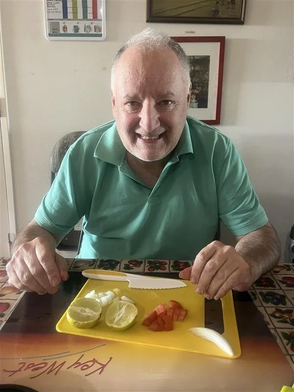 Smiling man sitting at a dining table with a yellow cutting board in front of him, containing lemon slices, chopped tomatoes, and a sliced onion, with a knife on the board.