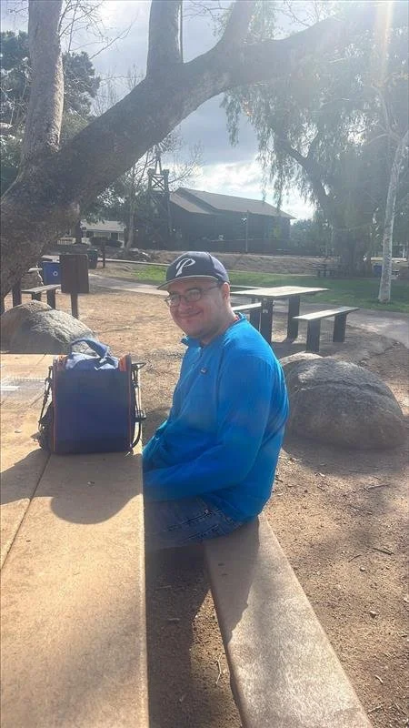 A man sitting on a park bench with a backpack beside him, smiling, with trees, a body of water, and a building in the background.