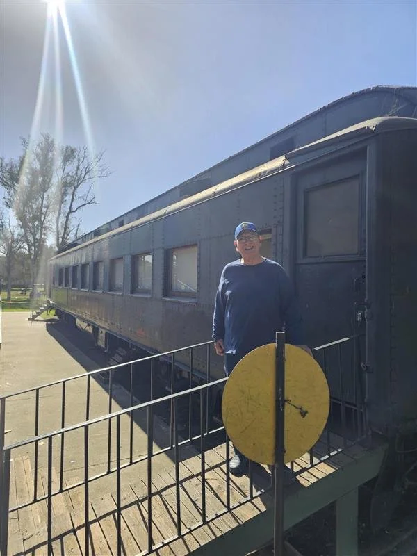 A person standing on a platform in front of a vintage train car, smiling at the camera with sunny weather and clear sky.