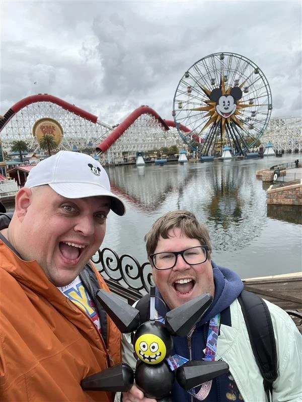 Two smiling men taking a selfie at an amusement park, with a large Mickey Mouse ferris wheel and a roller coaster in the background. One man is wearing an orange jacket and a white cap with a Mickey Mouse logo, and the other is wearing glasses, a dar