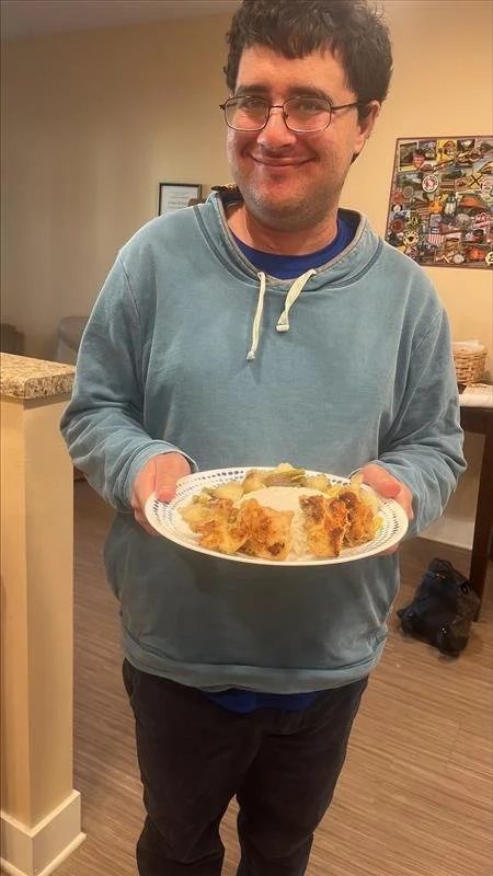 Young man with glasses in a blue hoodie smiling and holding a plate of baked casserole or pie in a home setting.