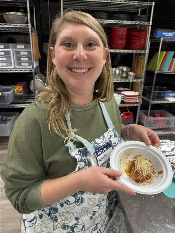 A woman with blonde hair smiling and holding a disposable plate with spaghetti and cheese in a kitchen or restaurant setting, wearing a green shirt and a patterned apron.