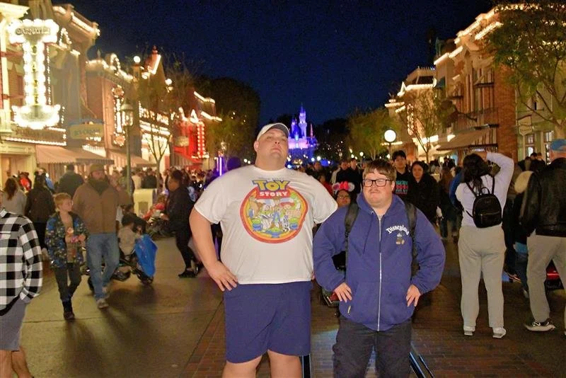 Two boys standing in front of a crowded Disney theme park street at night, with Main Street and a castle illuminated in the background, surrounded by visitors.