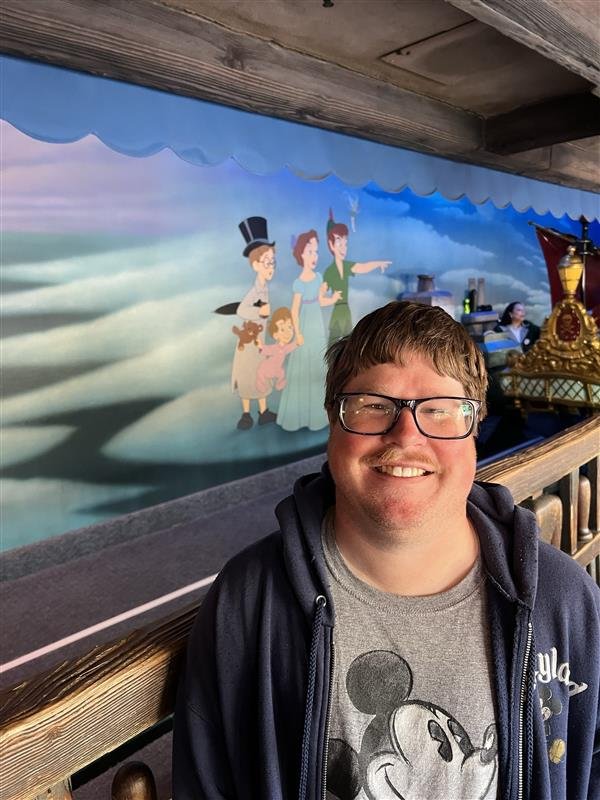 Smiling man with glasses in front of a mural depicting characters from Disney's Peter Pan, including Wendy, John, Michael, and Tinker Bell, in an indoor setting with a wooden railing and display of decorative items.