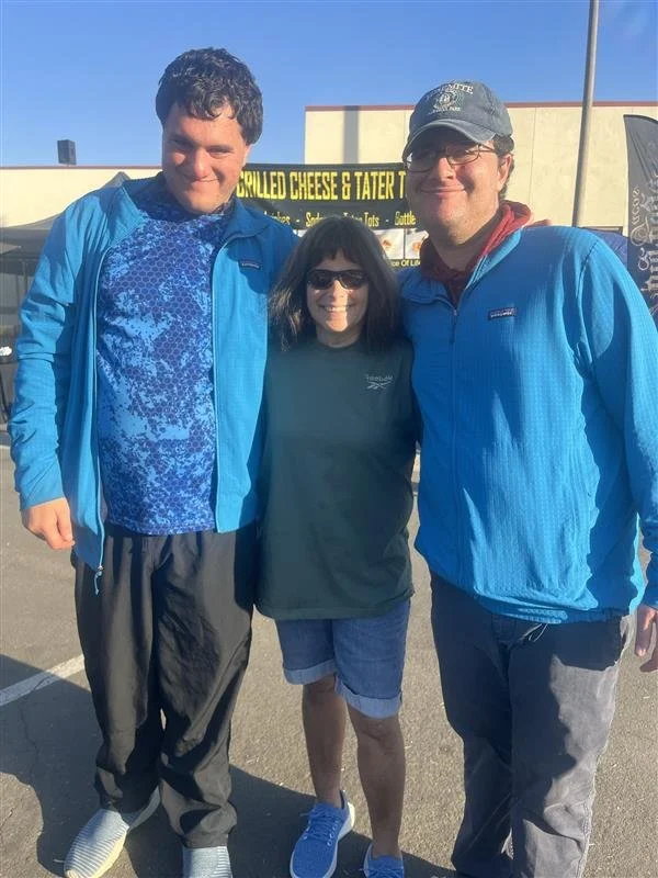 Three people standing together outdoors, smiling at the camera. Two men on either side of a woman, all dressed casually in jackets and shorts. There is a sign in the background indicating a food stand.