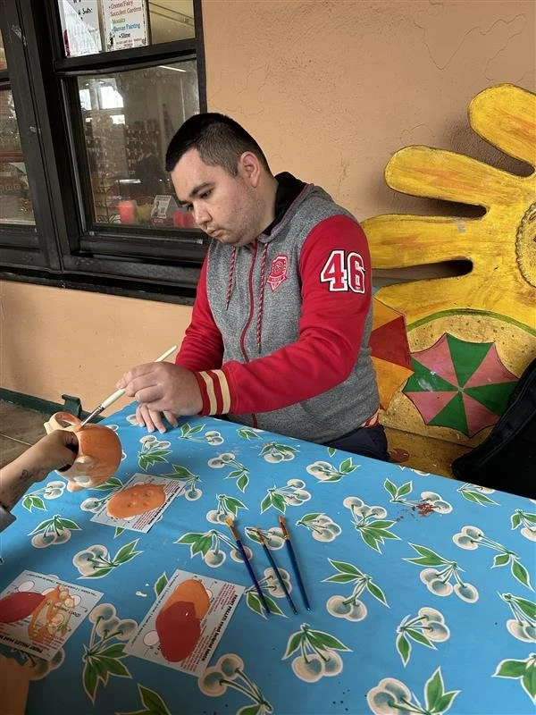 A young man with short dark hair is sitting at a table, focused on painting a ceramic object with a paintbrush. The table is covered with a blue tablecloth decorated with white and green floral designs. In the background, there is a window and colorf