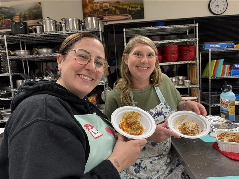 Two women smiling and holding plates of prepared food in a kitchen or food prep area with shelves of cookware and utensils in the background.