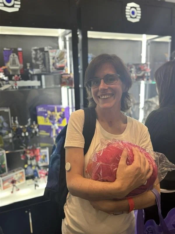 A woman with glasses, smiling, holding a pink item in a clear plastic bag, standing in front of a display case filled with toys, in a retail store.