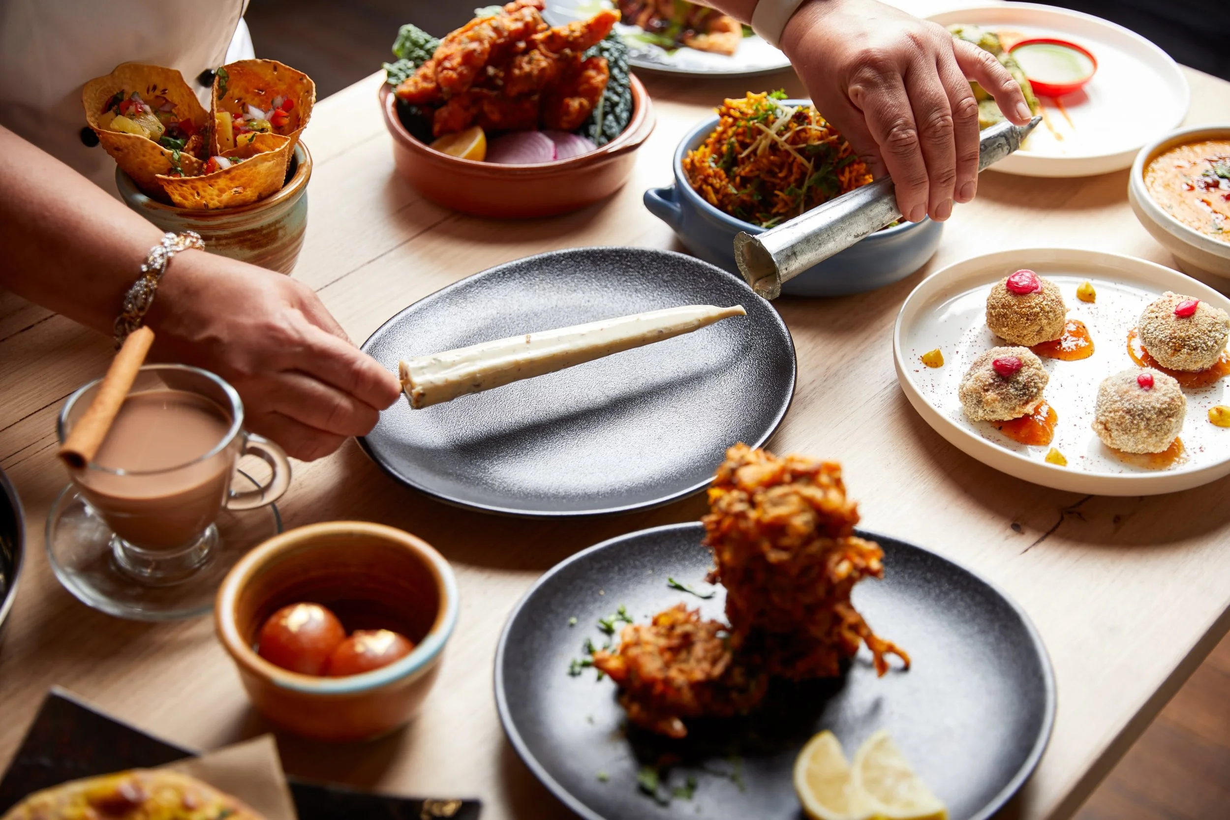 An Indian meal with various dishes including fried snacks, rice, curry, and desserts on a wooden table, with someone preparing food.