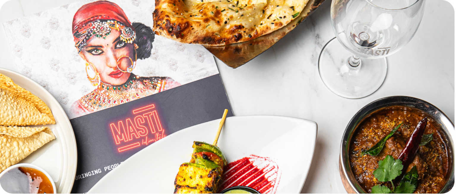 Assorted Indian dishes including naan bread, a bowl of curry, a skewer of grilled vegetables, and a glass of water on a white table, with a colorful booklet featuring a woman in traditional Indian attire.