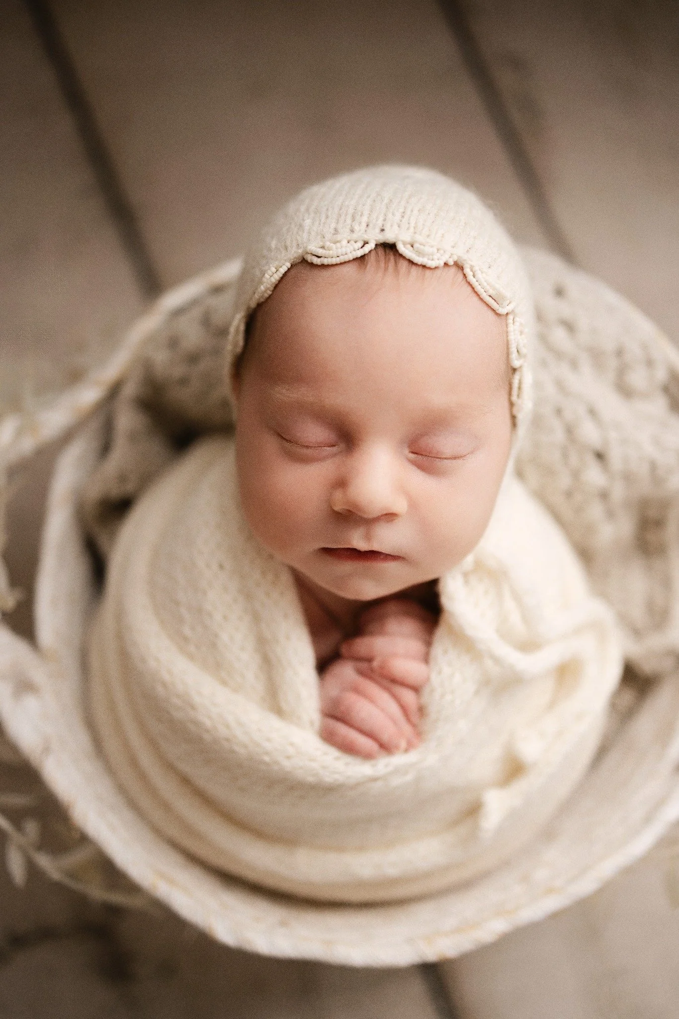 Newborn wearing a beaded cream bonnet with tiny hands tucked beneath her chin