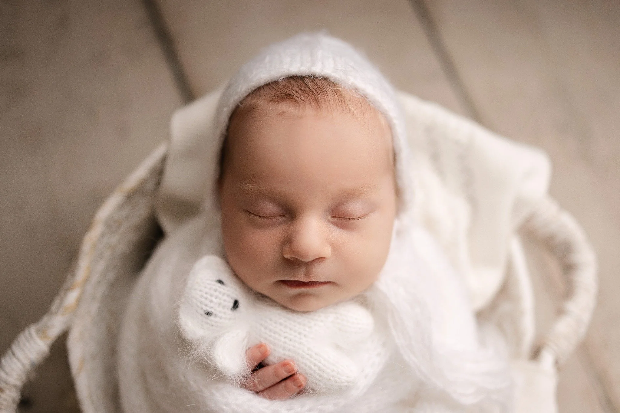 Close-up of a sleeping newborn in a soft cream bonnet holding a tiny knitted bear