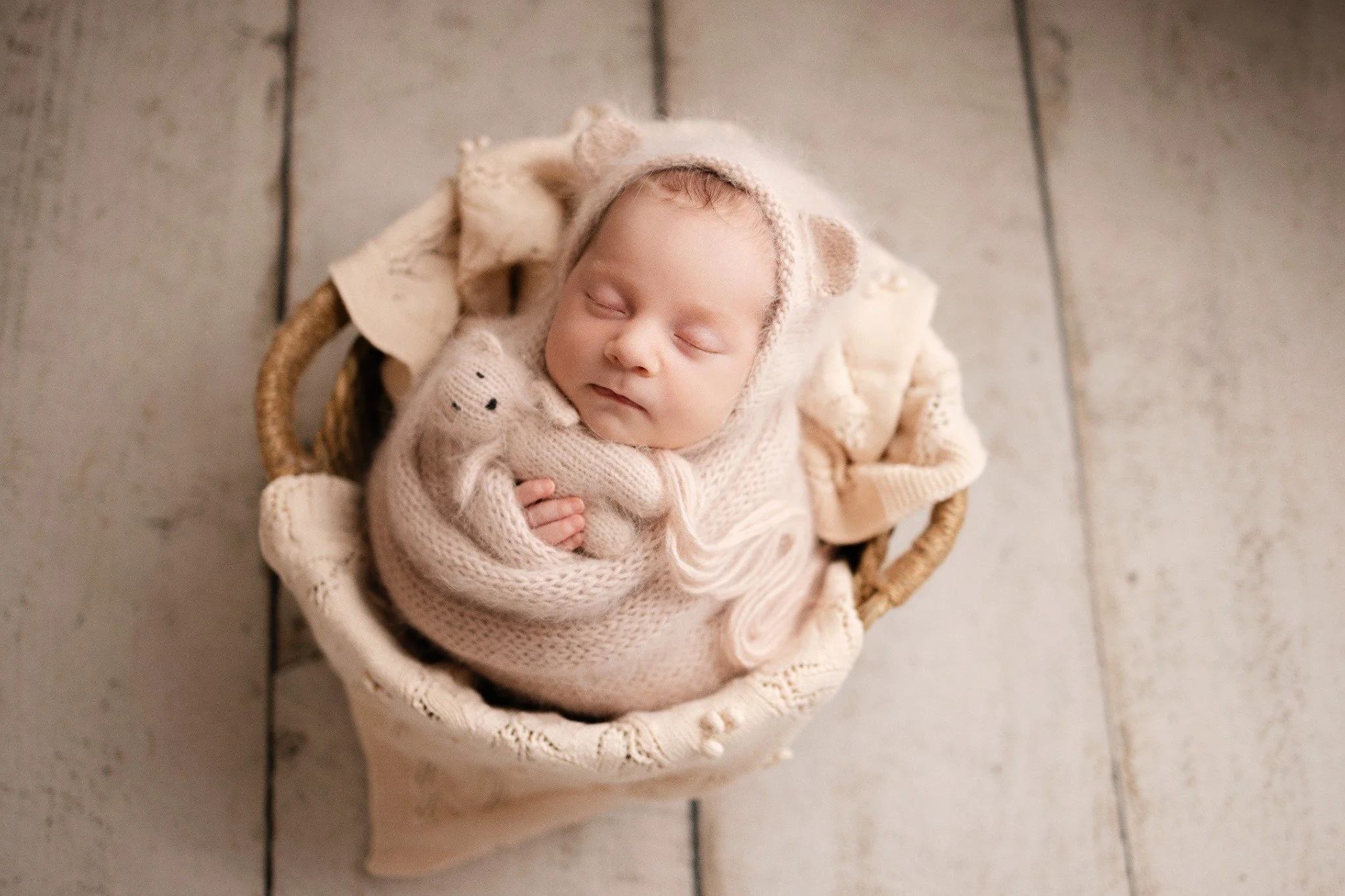 Newborn wrapped in oatmeal knit with a bear-ear bonnet and knitted bear in a wicker basket
