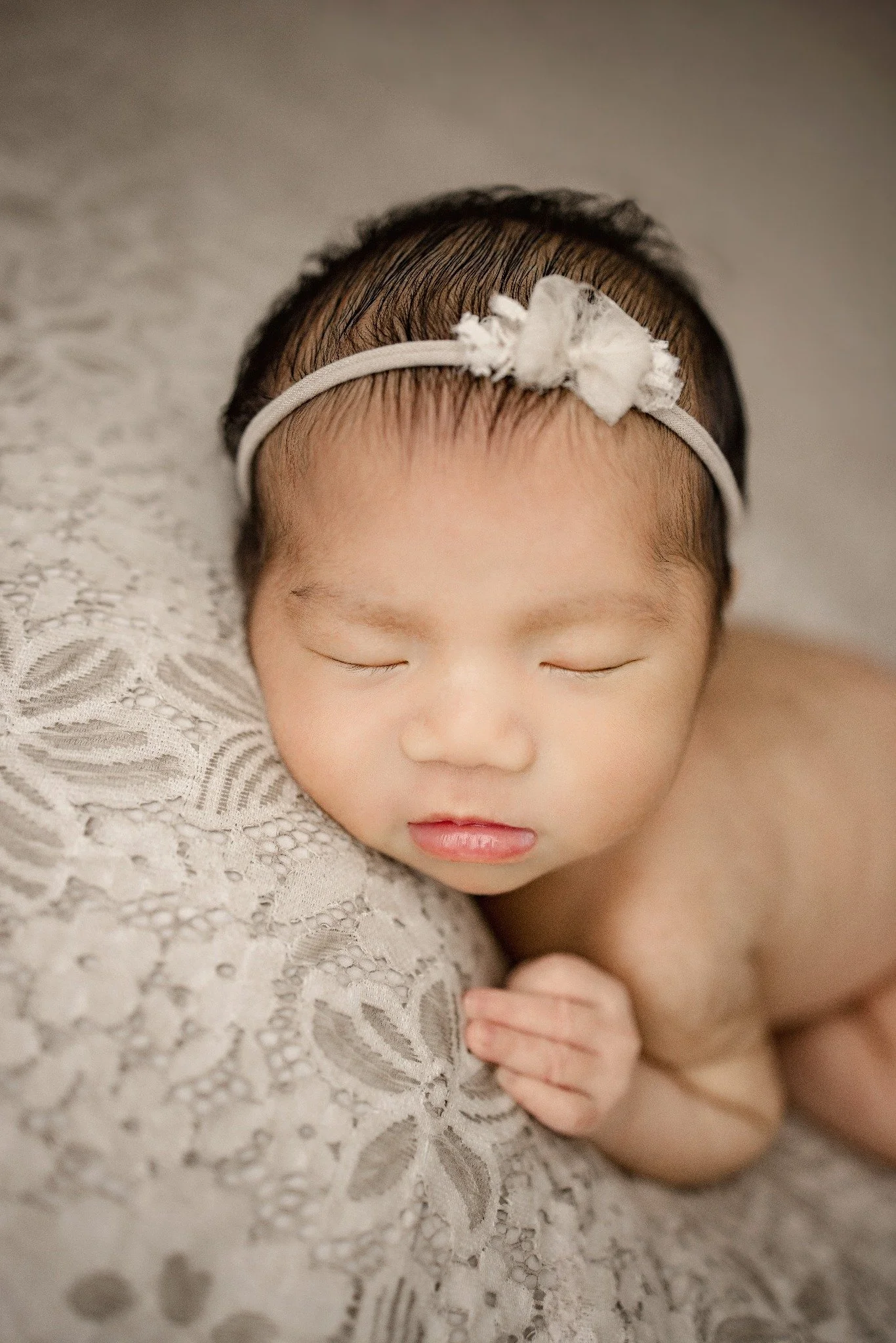 Close-up of a sleeping newborn wearing a cream floral headband, resting beside textured lace with one tiny hand visible.
