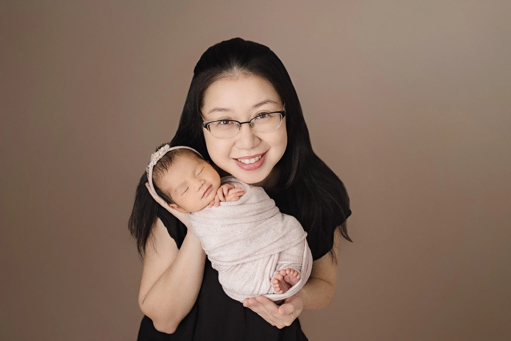 Mother holding her sleeping newborn in a pale wrap against a warm taupe backdrop.