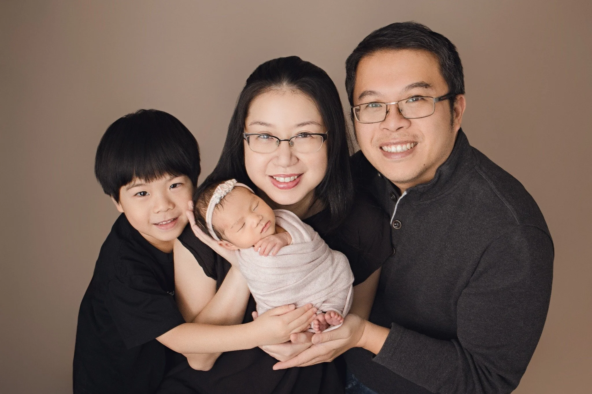 Parents and an older sibling gathered around a sleeping newborn in a pale wrap against a taupe backdrop.