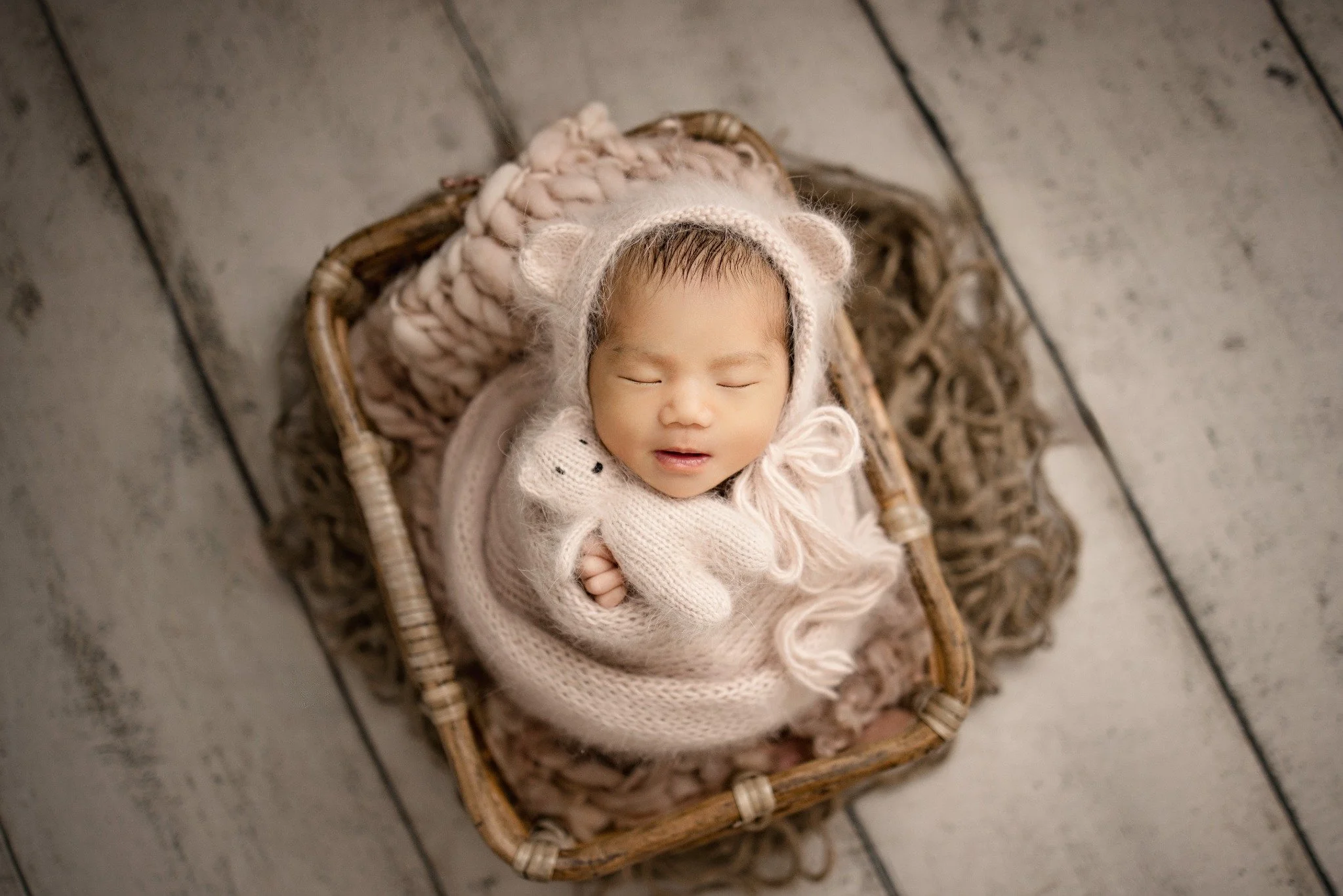 Sleeping newborn in a woven basket, wrapped in a pale knit blanket with a bear-ear bonnet and knitted toy.