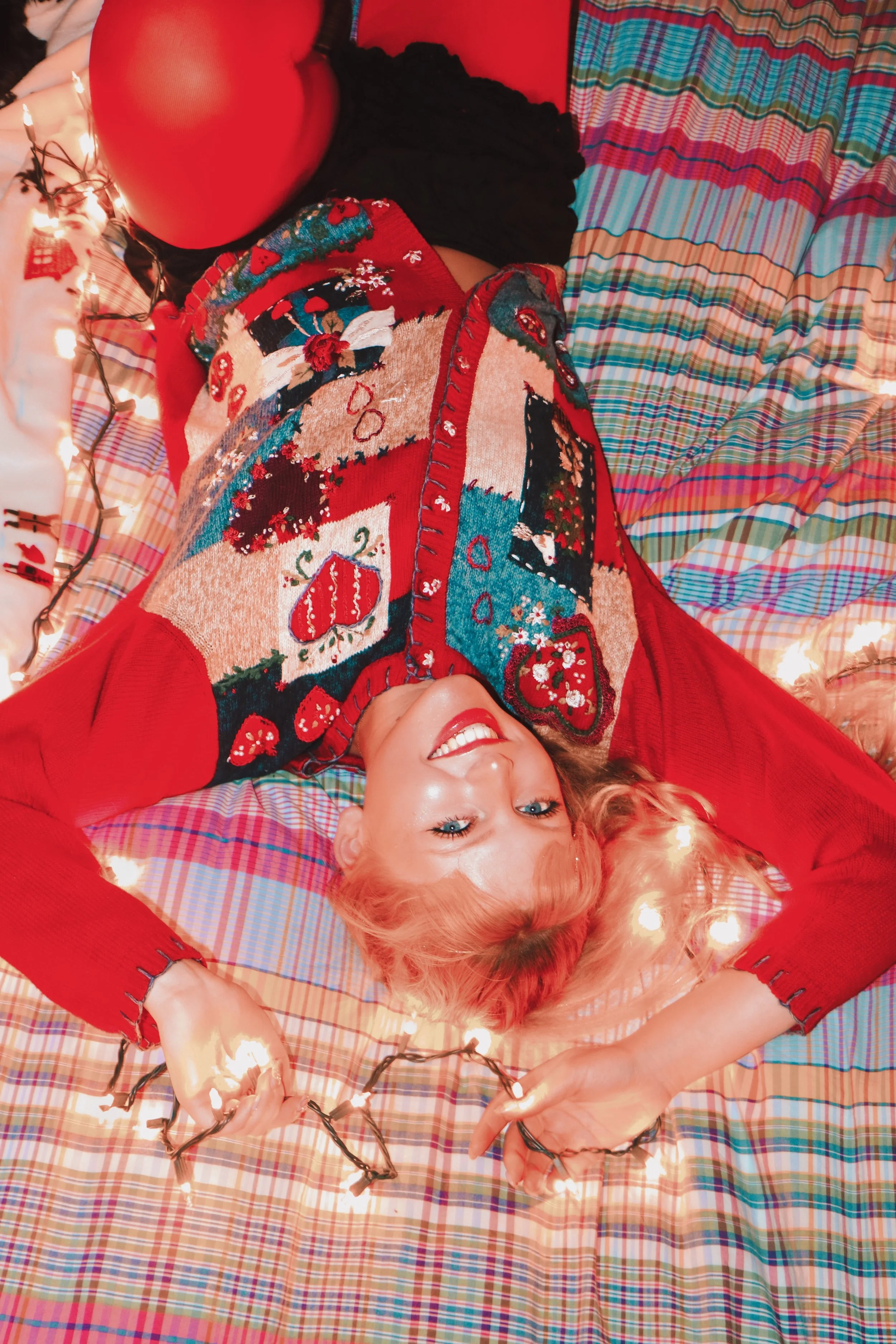 Person in red sweater with Christmas-themed design lying on a colorful plaid bedspread, smiling with string lights around them.