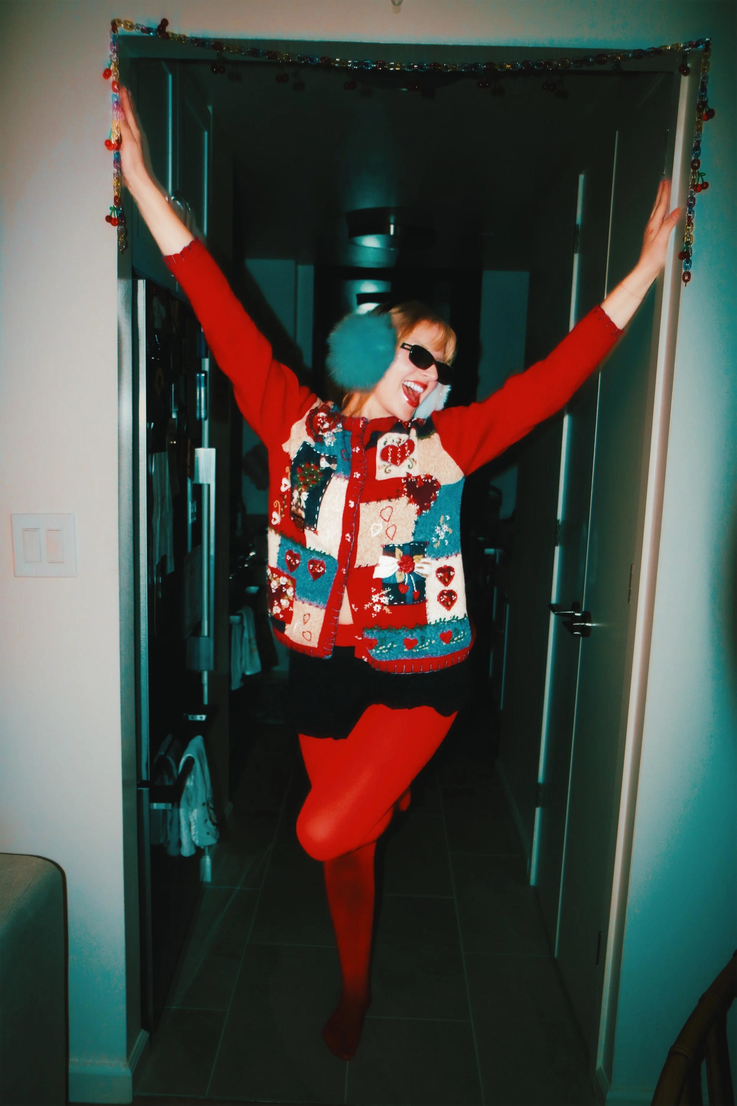 Person posing with arms raised, wearing colorful earmuffs, sunglasses, and a festive Christmas sweater in a doorway.