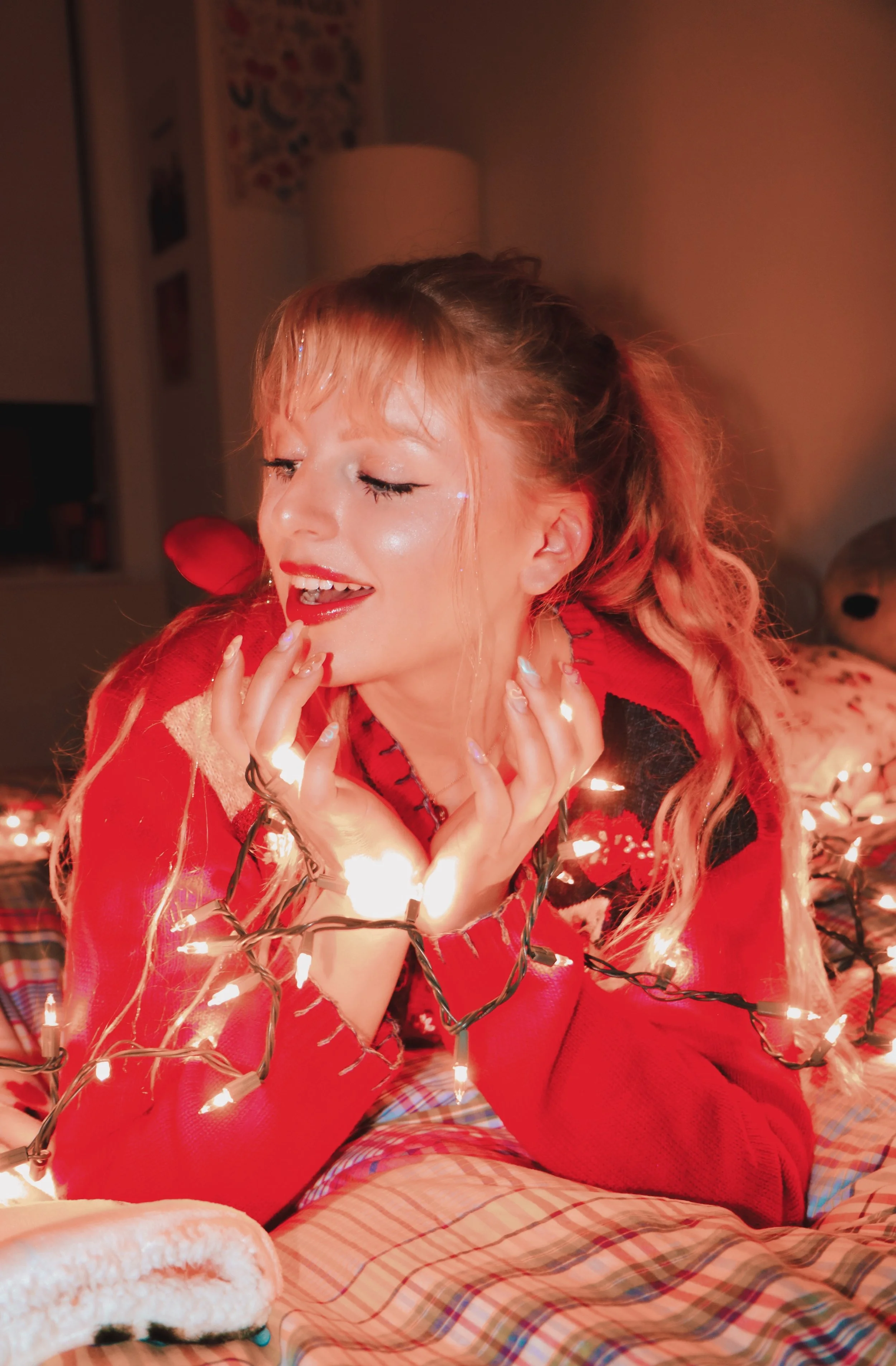 Person in a red sweater smiling while wrapped in Christmas lights, lying on a bed with a plaid blanket.