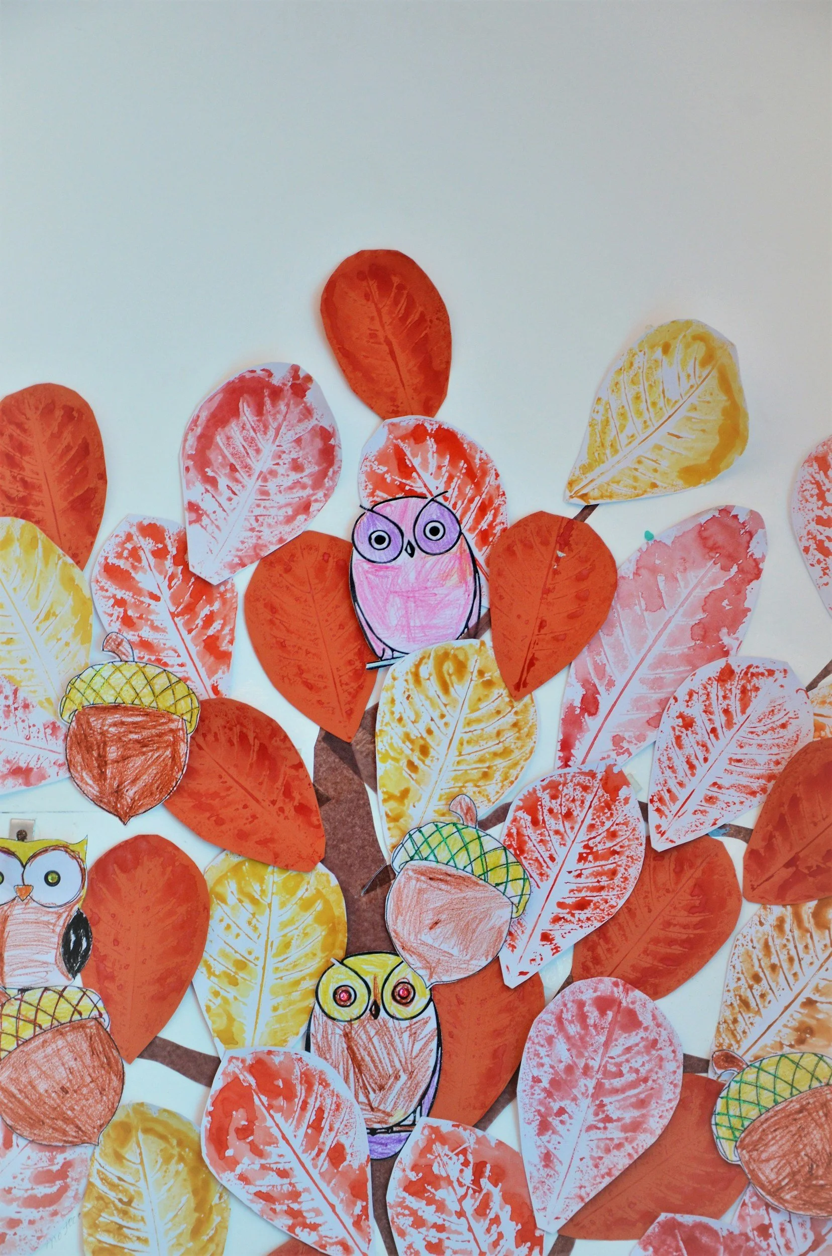 Children's craft art depicting trees made of paper leaves and cartoon owls and an owl drawn on paper, with leaves in shades of orange, yellow, and red.