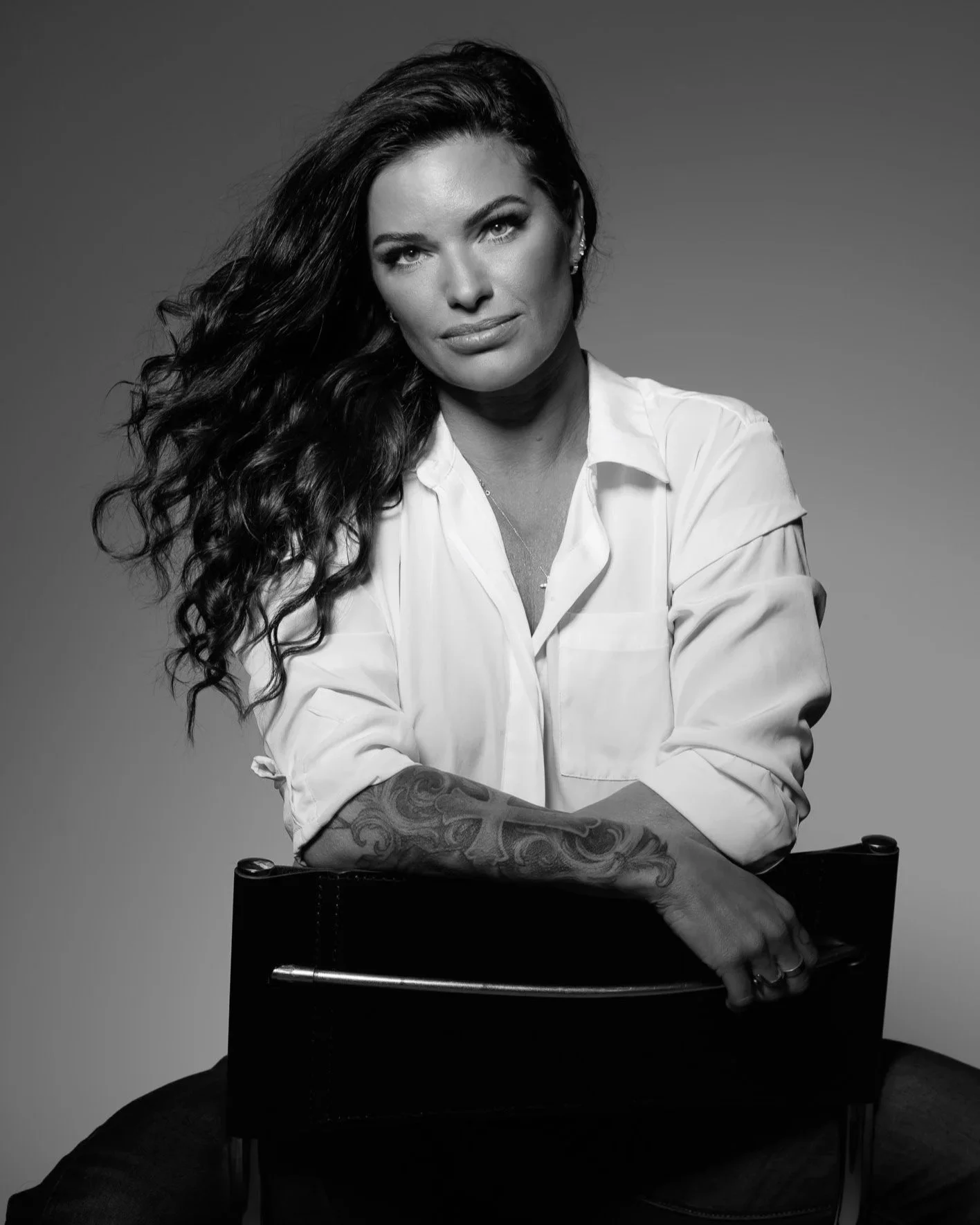 Black and white portrait of a woman with long curly hair, wearing a white shirt with rolled sleeves, sitting on a chair, looking directly at the camera with crossed arms resting on the chair's backrest.